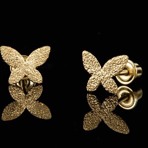 May include: A pair of gold butterfly earrings with a textured finish. The earrings are on a black background.