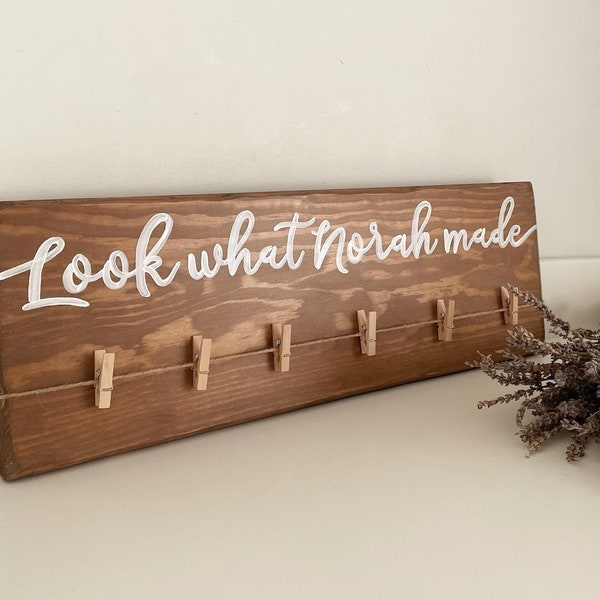 May include: A brown wooden sign with the text "Look what Norah made" written in white paint. The sign has a string with small wooden clothespins attached to it.