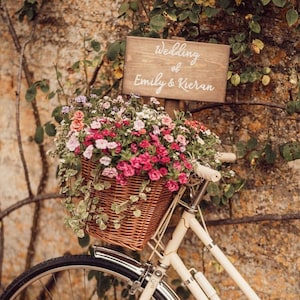 May include: A cream-coloured bicycle with a woven wicker basket filled with pink, purple, and white flowers. A rectangular wooden sign is attached to the handlebars, featuring the white script text Wedding of Emily & Kieran.