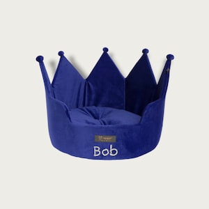 May include: A royal blue pet bed shaped like a crown. The bed has a soft, plush appearance with a rounded cushion inside. The name "Bob" is embroidered on the front. The bed has a tag that says "Hempster" on it.