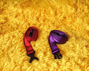 May include: Two rolled-up straps with black buckles. One strap is red with white dots, and the other is purple with white dots. The straps are on a yellow, fluffy surface.