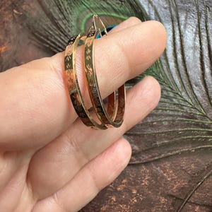 Puede incluir: Un par de pendientes de aro dorados con un patrón floral grabado repetitivo en la superficie exterior. Los aros tienen un acabado metálico pulido y un diseño circular fino, mostrando detalles grabados intrincados en la banda de metal.