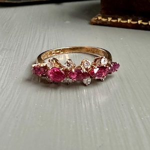 May include: A gold ring featuring a cluster of oval-cut, vibrant red gemstones, interspersed with smaller, clear stones. The ring's band is a warm gold color, and the gemstones are set in a delicate, ornate design. This piece is a statement jewelry item.
