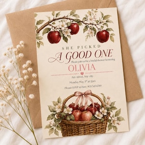 May include: A cream-colored bridal shower invitation card featuring a watercolor illustration of apple branches at the top and a woven basket filled with red apples at the bottom. The text reads: SHE PICKED A GOOD ONE, Please join us for a bridal shower honoring OLIVIA, Any street, Any city, Monday May 3rd at 2pm, Please RSVP to Kim 905 345 6789.