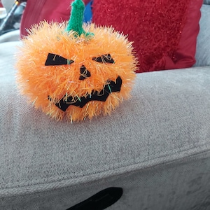 Può includere: Una zucca arancione pelosa con una faccia di zucca intagliata nera, seduta su un divano grigio.