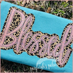 May include: A turquoise blue t-shirt with a pink and brown "Blessed" graphic with a leopard print design. The graphic is outlined in pink and has a glittery finish.