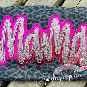 Puede incluir: Un diseño de palabra "Mama" rosa y plateado sobre una tela de estampado de leopardo negro y blanco.