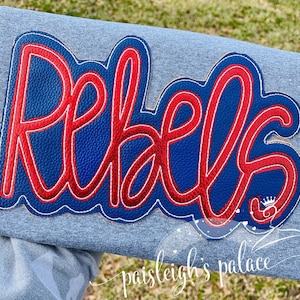 May include: Red and blue felt applique with the word "Rebels" in a cursive font. The applique is on a gray t-shirt.