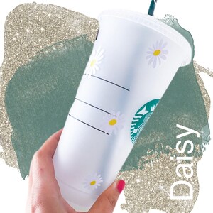 May include: White reusable cup with a black stripe and daisy decals. The cup has a green Starbucks logo and the word "Daisy" written in black.