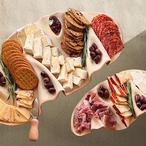May include: Two cream-coloured, wavy-edged ceramic serving platters arranged with assorted charcuterie, including sliced salami, prosciutto, various cheeses, crackers, dark olives, sprigs of rosemary, a small dollop of jam, and a metal serving knife.