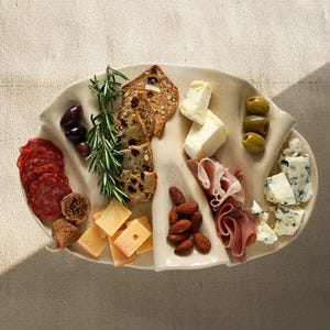 May include: A charcuterie board with an assortment of food items. Sliced salami, figs, cheese cubes, almonds, prosciutto, olives, and crackers are arranged on a cream-colored, leaf-shaped platter. Rosemary sprigs add a fresh touch.