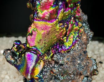 May include: A close-up of a mineral specimen with a vibrant, iridescent surface. The mineral displays a spectrum of colors, including pink, green, yellow, and blue, with a rough, textured appearance. The mineral sits on a bed of small, white crystals.
