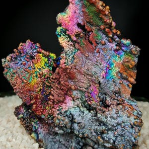 Puede incluir: Primer plano de un espécimen mineral con una superficie vibrante e iridiscente. La roca muestra un espectro de colores, incluyendo azules, verdes, rosas y naranjas, con una apariencia texturizada y con bultos. El mineral se encuentra sobre un lecho de pequeñas piedras blancas granulares.