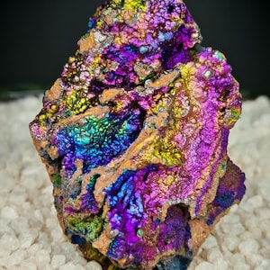 Op de afbeelding: Een levendig, iriserend mineraal specimen met een ruw, getextureerd oppervlak. De steen toont een spectrum aan kleuren, waaronder paars, blauw, groen en goud. Het mineraal ligt op een bed van kleine, witte, kristallijne rotsen.