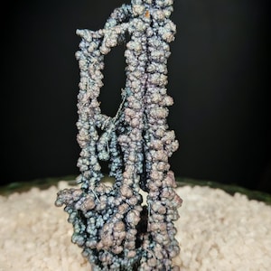 Könnte beinhalten: Einzigartiges, schillerndes Mineralexemplar mit strukturierter, blasenartiger Oberfläche. Das Mineral zeigt Farbtöne von Blau, Lila und Silber und ist vor dem Hintergrund weißer Kieselsteine und einem dunklen Hintergrund platziert. Die Gesamtform ist vertikal und organisch.