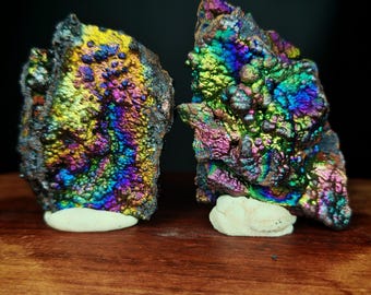 May include: Two iridescent mineral specimens with vibrant colors of blue, green, yellow, and purple. The minerals sit on small, white bases, displayed against a dark background. The rough texture and play of colors create a striking visual.