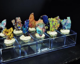 May include: A collection of iridescent mineral specimens in various colors, including blue, orange, and green, displayed on individual white bases within a clear acrylic display case. The minerals have a textured, crystalline appearance.