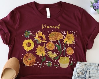 May include: A maroon t-shirt with a sunflower and flower design and the word "Vincent" printed on the front. The shirt is laid flat on a white surface with a pair of brown wedges and a pair of denim shorts.