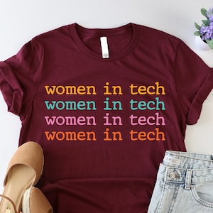 May include: A maroon t-shirt with the text "women in tech" repeated four times in different colors. The text is in a colorful font. The shirt is laid flat on a white surface with a pair of brown wedges and denim shorts.