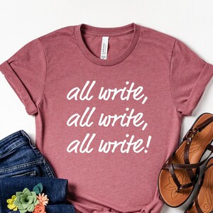 May include: A mauve t-shirt with the text "all write, all write, all write!" in white lettering. The shirt is paired with blue jeans and brown sandals.