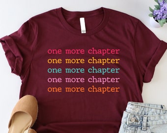 May include: A maroon t-shirt with the text "one more chapter" repeated five times in different colors. The text is in a rainbow color scheme, with each line of text in a different color.