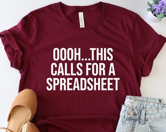 May include: A maroon t-shirt with white text that reads "OOOH... THIS CALLS FOR A SPREADSHEET". The shirt is laid flat on a white surface with a pair of brown sandals and denim shorts.