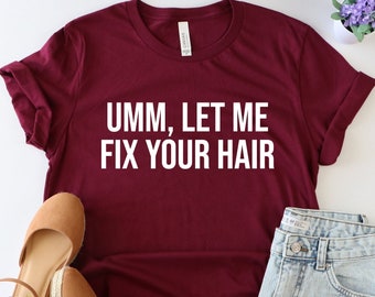 May include: Burgundy t-shirt with white text that reads "Umm, let me fix your hair". The shirt is laid flat on a white surface with a pair of brown sandals, denim shorts, and a small potted plant with purple flowers.