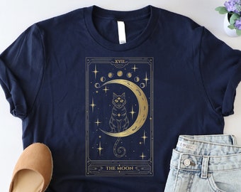 May include: A navy blue t-shirt with a gold tarot card design featuring a cat sitting on a crescent moon. The card reads "XVIII - THE MOON".