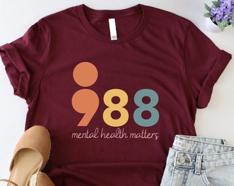Könnte beinhalten: Ein maroonfarbenes T-Shirt mit einer bunten Grafik eines Semikolons und der Zahl 988. Der Text "mental health matters" ist unter der Grafik aufgedruckt.