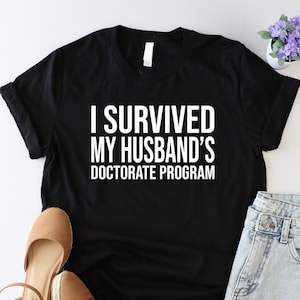 May include: Black t-shirt with white text that reads "I survived my husband's doctorate program". The shirt is laying flat on a white surface with a pair of brown sandals and denim shorts.