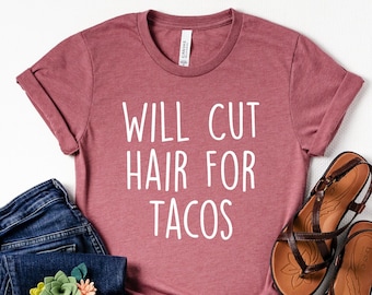 May include: A maroon t-shirt with the text "WILL CUT HAIR FOR TACOS" in white, paired with blue jeans and brown sandals. The jeans have a floral embellishment. The sandals are leather with multiple straps.