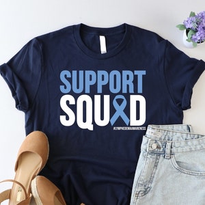 May include: A navy blue t-shirt with white text that reads "SUPPORT SQUAD" with a blue ribbon in the middle of the word "SQUAD". The text "KYPHOSIS AWARENESS" is written in smaller white letters below the ribbon. The shirt is laid flat on a white surface with a pair of brown wedges and denim shorts.