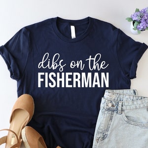 May include: A navy blue t-shirt with the text "dibs on the fisherman" in white lettering. The shirt is paired with denim shorts and brown wedges.