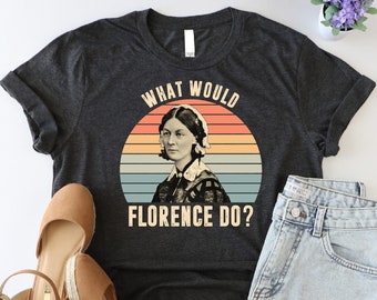 May include: A dark gray t-shirt with a vintage-style graphic of Florence Nightingale. The graphic features a sunset-colored circle with Florence Nightingale's portrait in the center. The text "What Would Florence Do?" is printed below the graphic.