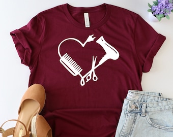 May include: A maroon t-shirt with a white heart design featuring a hair dryer, scissors, and a comb. The shirt is laid flat on a white surface with a pair of brown wedges and denim shorts.