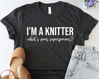 May include: A dark gray t-shirt with the text "I'm a knitter what's your superpower?" in white. The shirt is paired with denim shorts and brown wedges.