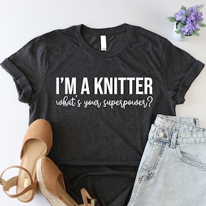 May include: A dark gray t-shirt with the text "I'm a knitter what's your superpower?" in white. The shirt is paired with denim shorts and brown wedges.