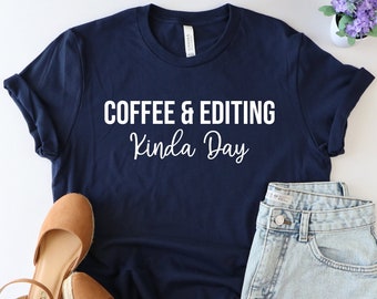 May include: A navy blue t-shirt with white text that reads "Coffee & Editing Kinda Day". The shirt is paired with denim shorts and brown sandals.
