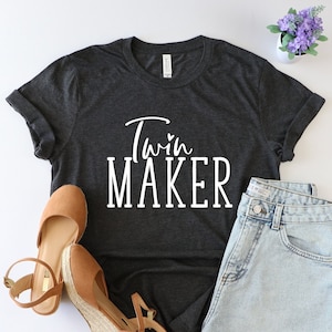 May include: A dark gray t-shirt with the text "Twin Maker" in white lettering. The shirt is paired with denim shorts and brown wedges.