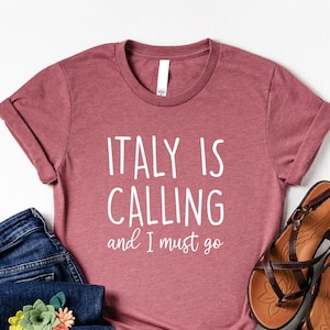 May include: A maroon t-shirt with the text "ITALY IS CALLING and I must go" in white lettering. The shirt is paired with blue jeans and brown sandals.