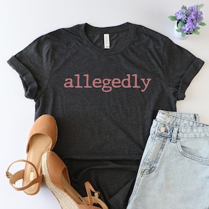 Op de afbeelding: Een donkergrijs T-shirt met het woord "allegedly" in roze op de voorkant. Het shirt is gestyled met een spijkerbroekje en bruine wedges.