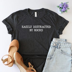 May include: A dark gray t-shirt with the text "EASILY DISTRACTED BY ROCKS" in white. The shirt is laid flat on a white surface with a pair of brown wedge sandals, a pair of denim shorts, and a small potted plant with purple flowers.