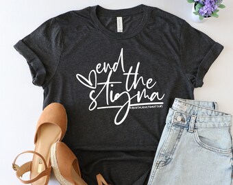 May include: A dark grey t-shirt with the text "end the stigma" in white lettering. The shirt is styled with denim shorts and brown wedges.