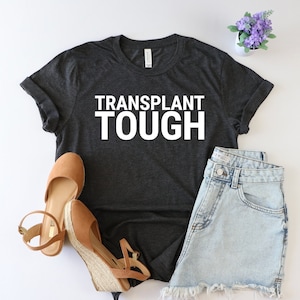 May include: A dark gray t-shirt with the text "TRANSPLANT TOUGH" in white lettering. The shirt is laid flat on a white surface with a pair of brown sandals and a pair of light blue denim shorts.