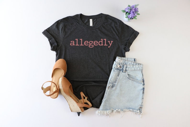 Op de afbeelding: Een donkergrijs T-shirt met het woord "allegedly" in roze op de voorkant. Het shirt is gestyled met een spijkerbroekje en bruine wedges.