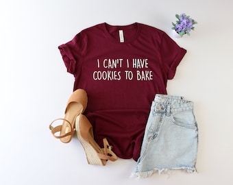 May include: A maroon t-shirt with the text "I can't I have cookies to bake" in white lettering. The shirt is laid flat on a white surface with a pair of brown sandals and denim shorts.