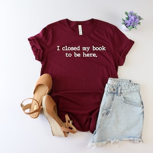 May include: Burgundy t-shirt with white text that reads "I closed my book to be here."