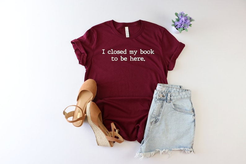 May include: Burgundy t-shirt with white text that reads "I closed my book to be here."