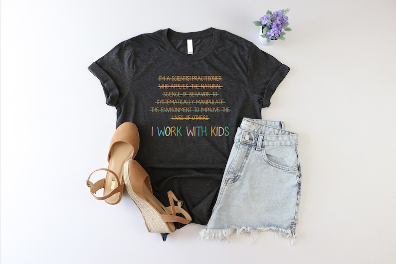 K&ouml;nnte beinhalten: Ein dunkelgraues T-Shirt mit einer humorvollen Botschaft &uuml;ber die Arbeit mit Kindern. Der Text lautet: "Ich bin ein Wissenschaftler/Praktiker, der die Naturwissenschaften des Verhaltens anwendet, um die Umwelt systematisch zu manipulieren, um das Leben anderer zu verbessern. Ich arbeite mit Kindern."