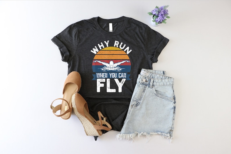 May include: A dark grey t-shirt with a vintage sunset graphic and the text "Why run when you can fly". The graphic features a swimmer in a blue silhouette.